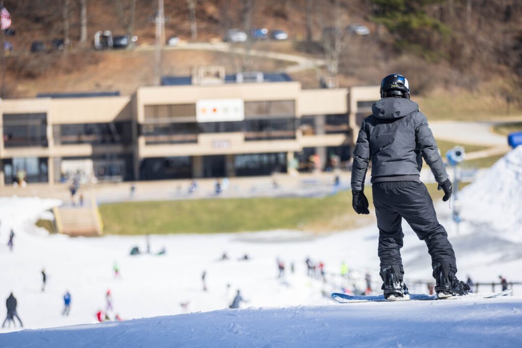 A person in a navy blue snow suit and helmet standing at the top of a snowy slope on a snowboard.