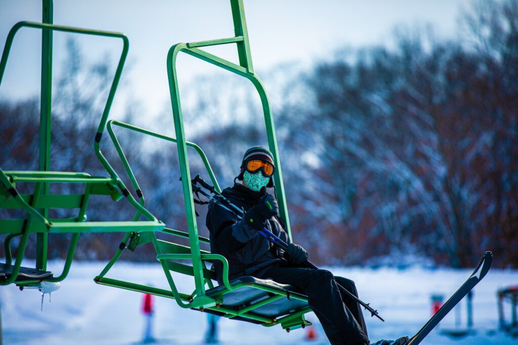 A person dressed in ski apparel riding a green ski lift chair and giving a thumbs up sign to the camera.