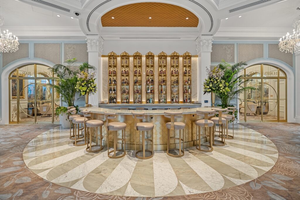 A gold and ivory commercial round bar with cream colored leather bar stools in an open area between arched glass doors trimmed in gold surrounded by marble flooring by Armina Stone.