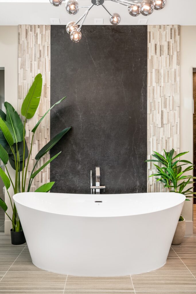 A white bathtub with a gold faucet sits in front of a black stripe in the bathroom wall, surrounded by plants. 