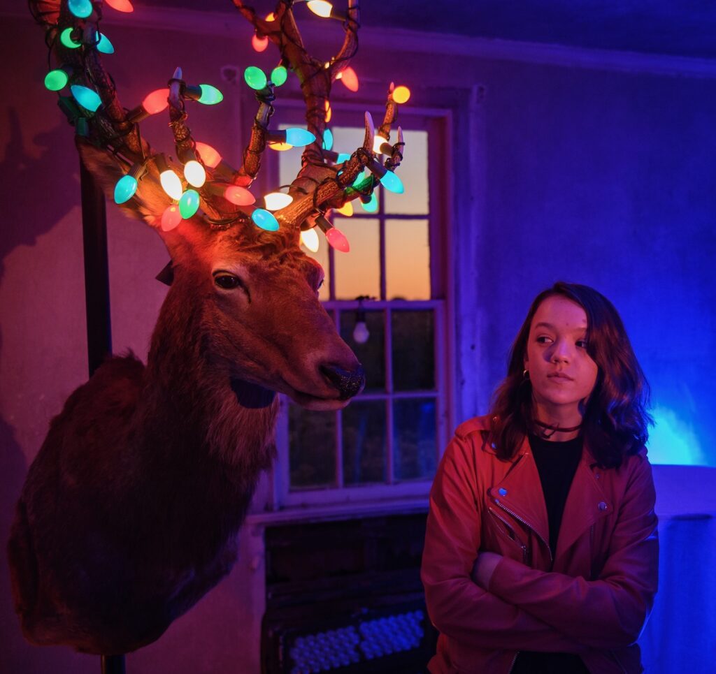 A woman stands beside a deer head with Christmas lights on it, in a purple-lit room