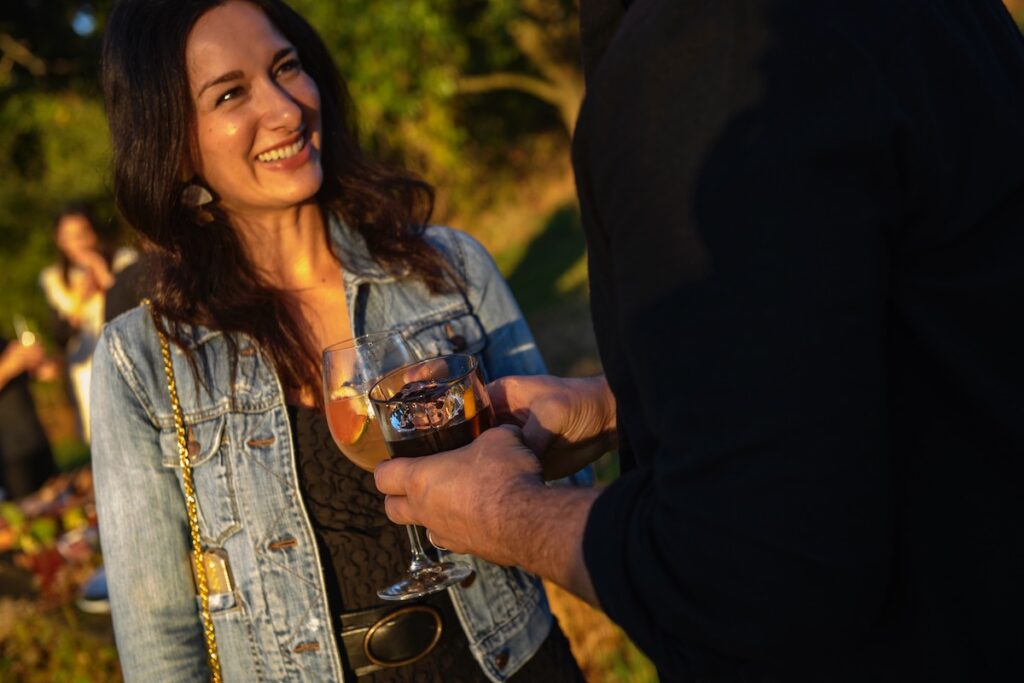 A woman in a denim jacket stands with a glass of wine at an outdoor party