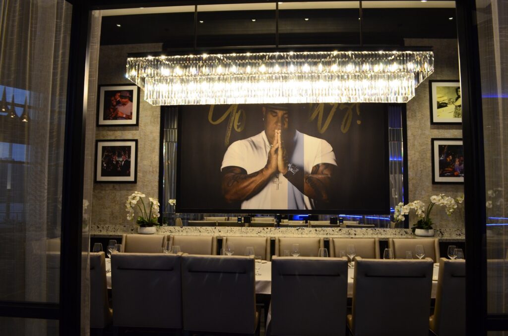 A long dining table at Rivers Casino with a rectangular chandelier above, sitting in front of a backdrop of a man in a white short sleeved shirt with tattooed arms and his hands pressed together in front of his face.