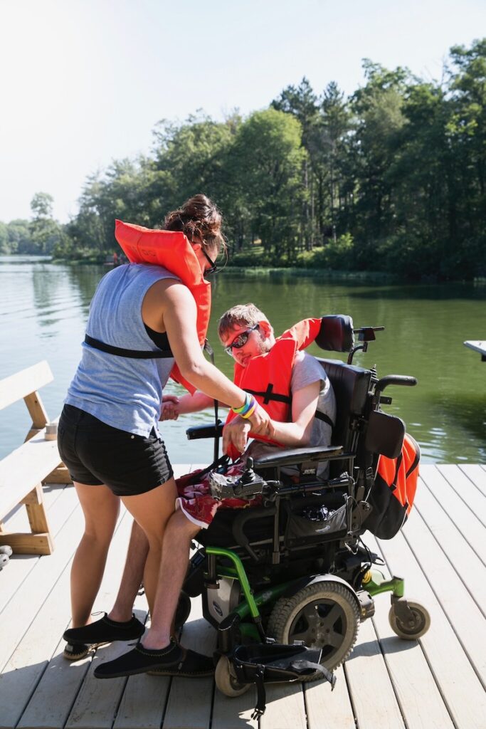 A woman in a life vest helps a boy in a wheelchair get his life vest on while on a wooden dock.