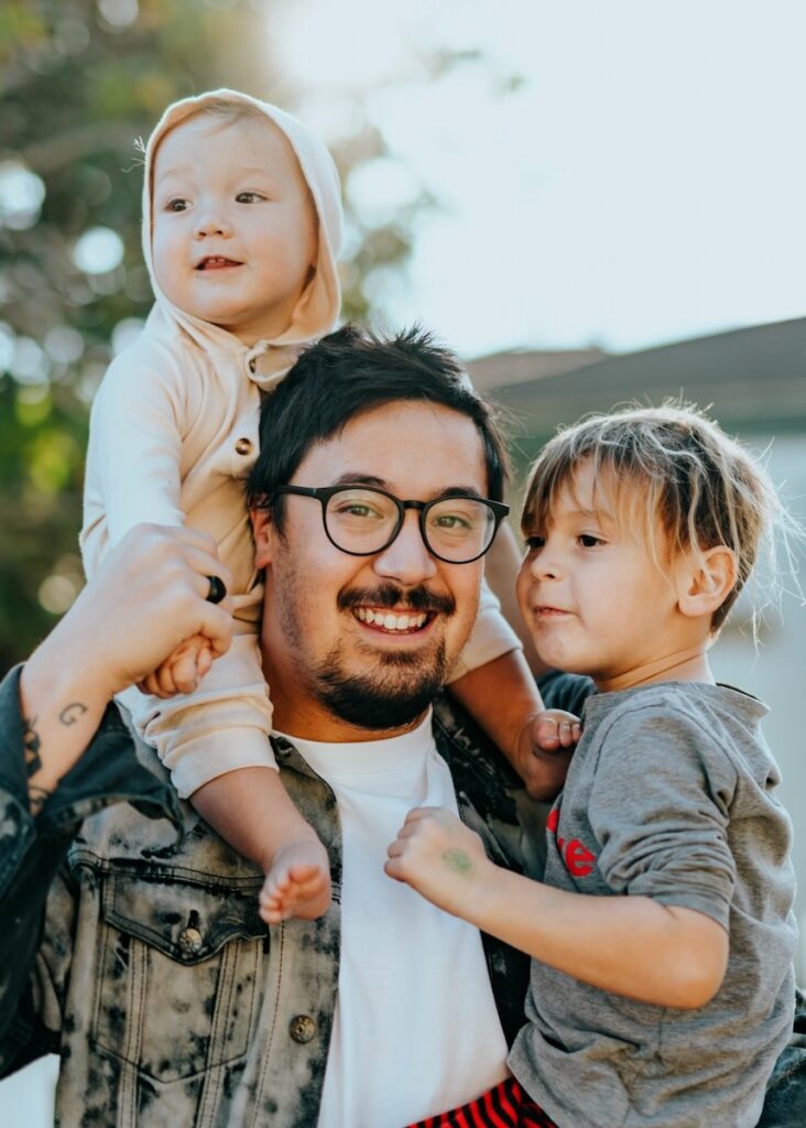 A dad with a small son on his back and other in his arms.