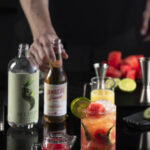 A white man's hand hold a bottle of ginger beer as it sits on a black table. Next to the soda sits a mojito mocktail orange in color.