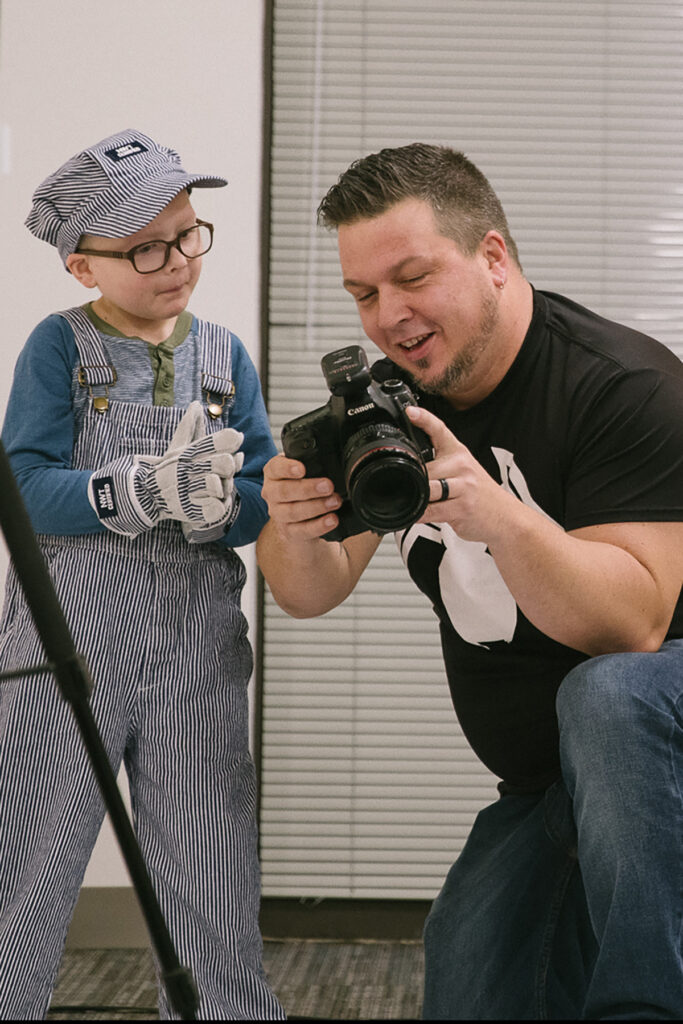 A man with a camera shows it to a young boy wearing a train conductor's uniform.