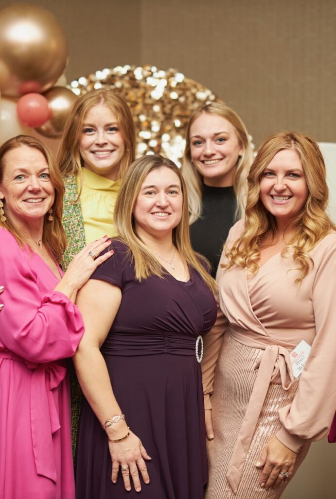 A group of 5 women in dresses stand in front of gold celebratory decor.