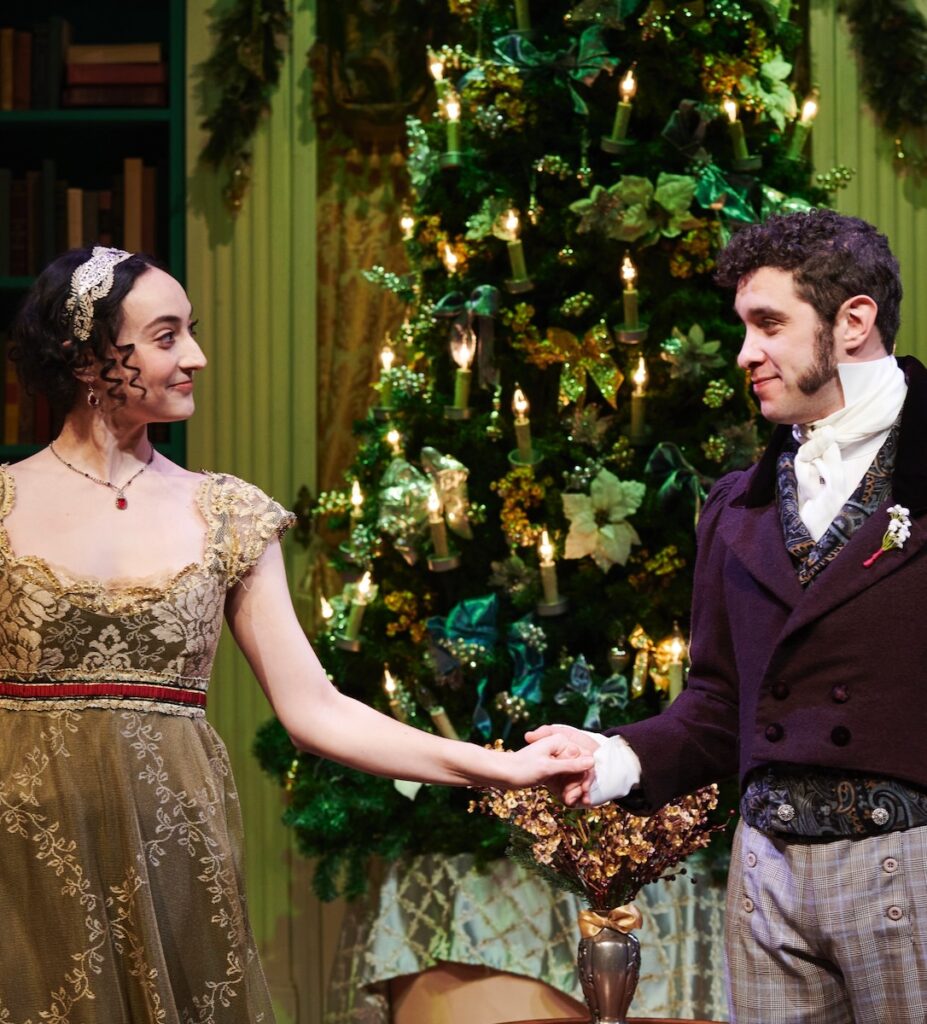 A woman in a dress and man in a dress coat take each other's hand in front of a Christmas tree with lights on it.