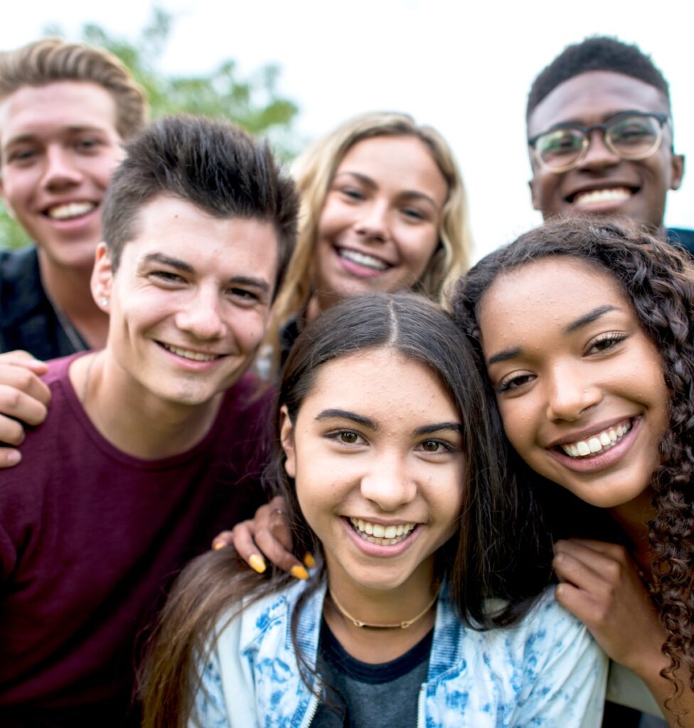 A multi-ethnic group of teenagers are outdoors on a cloudy day. They are wearing casual clothing. They are smiling while taking a selfie together.