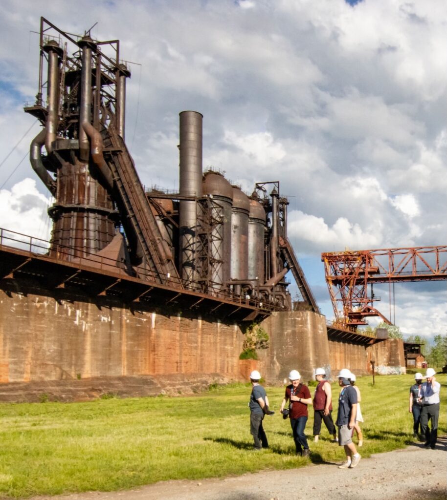 A large steel mill building sits outside while a group of people in white construction helmets walk around the ground.