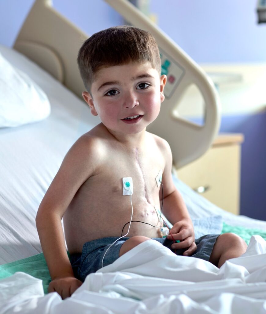 A small boy without a shirt on sits in a hospital bed with a blanket over him.