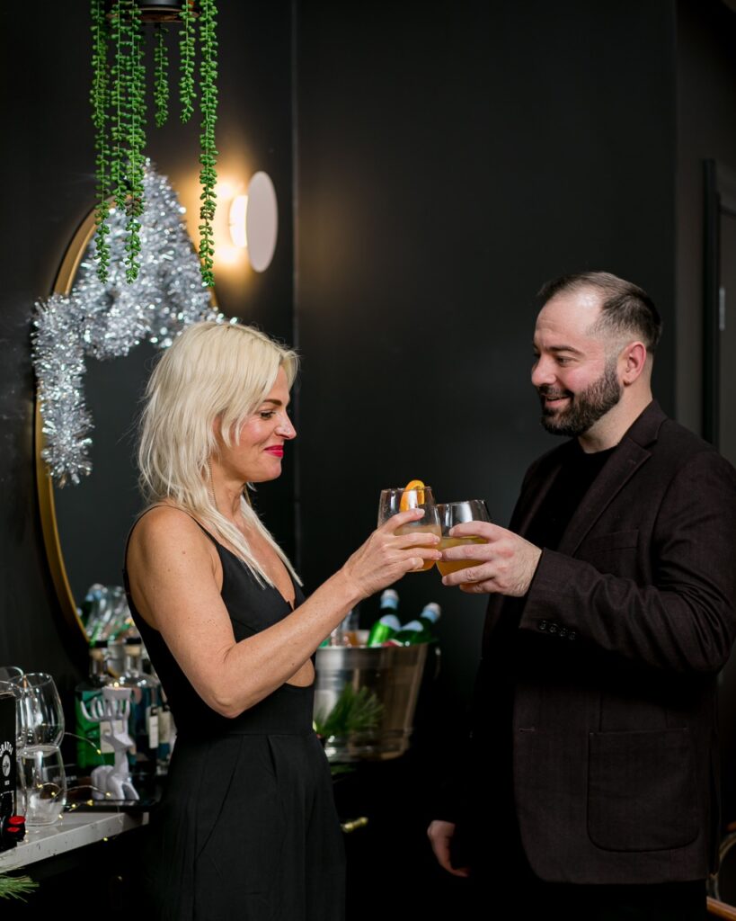 Two workers, a man and woman, in black clothing clink together their orange cocktail glasses at a holiday office party.