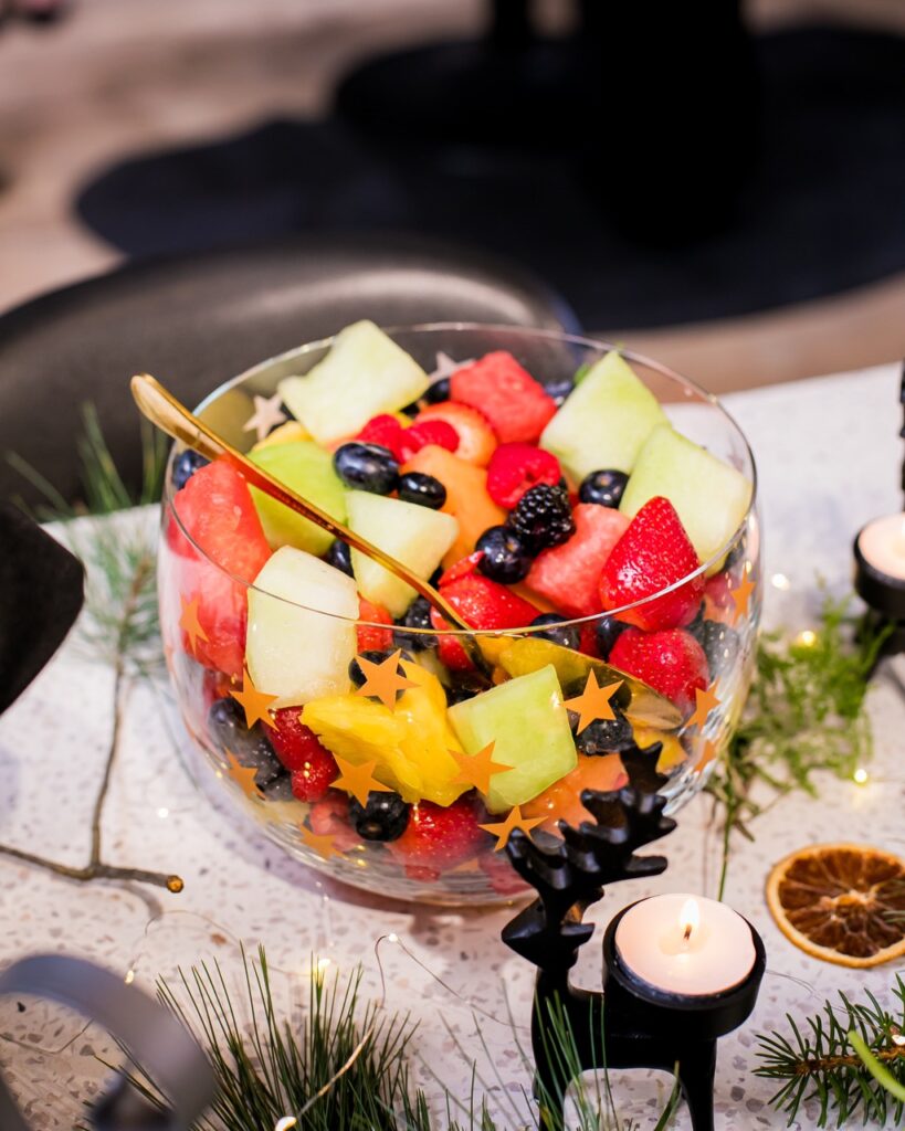 clear glass bowl holds a fruit salad with melon, cantaloupe, strawberries, and blueberries on a white table cloth.