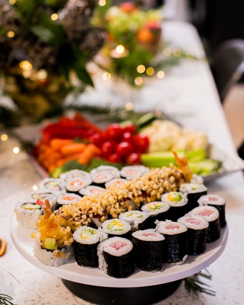 A white place holds an arrangement of 5 different sushi types, each in a line and all sitting amongst a white table with a veggie tray on it.