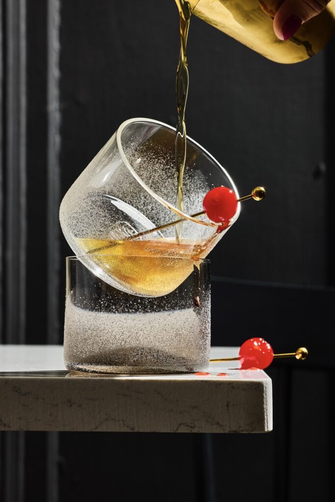 A short glass with bourbon being poured into it as a toothpick with a cherry hangs out of the glass.