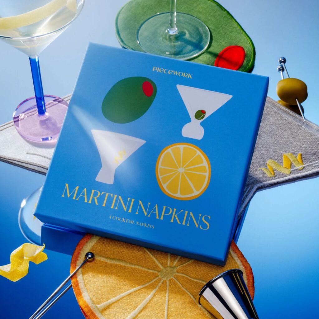 A martini napkins set sits on a blue table with a martini glass, olive, and orange coasters.