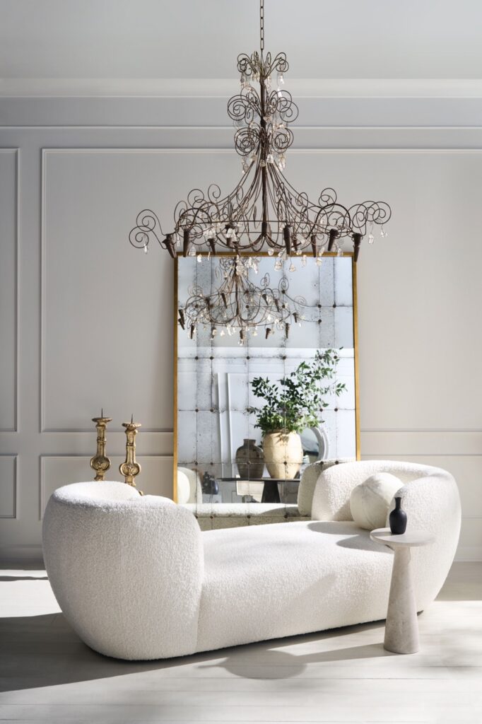 A white chaise sits in front of a large mirror and below a chandelier.