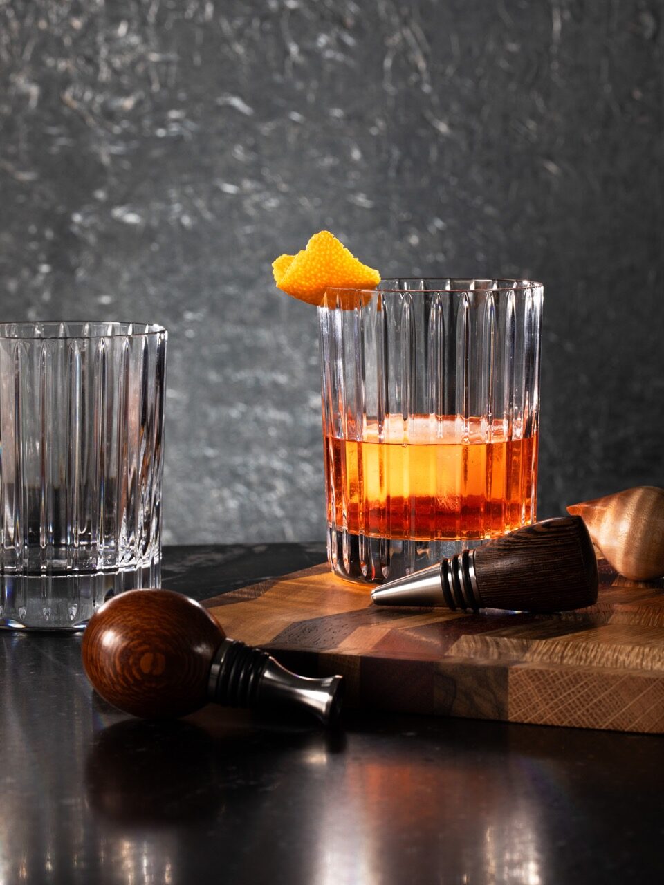 A cocktail glass with an orange drink instead and orange garnish sits behind two wood top wine stoppers as another empty glass sits off to the left.