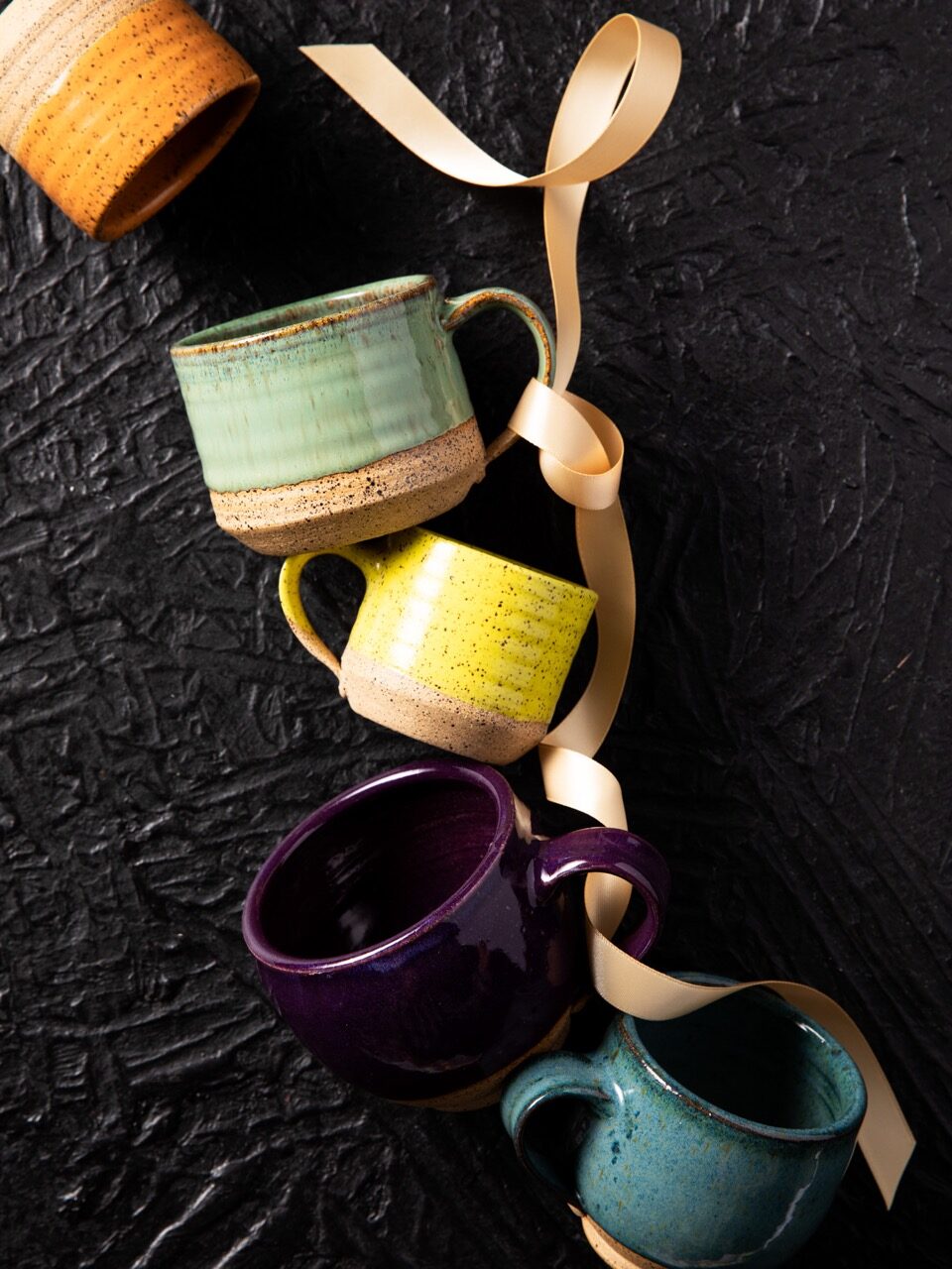 An orange, green, yellow, purple, and blue mugs sit in a line, their handles laced through a pale ribbon.