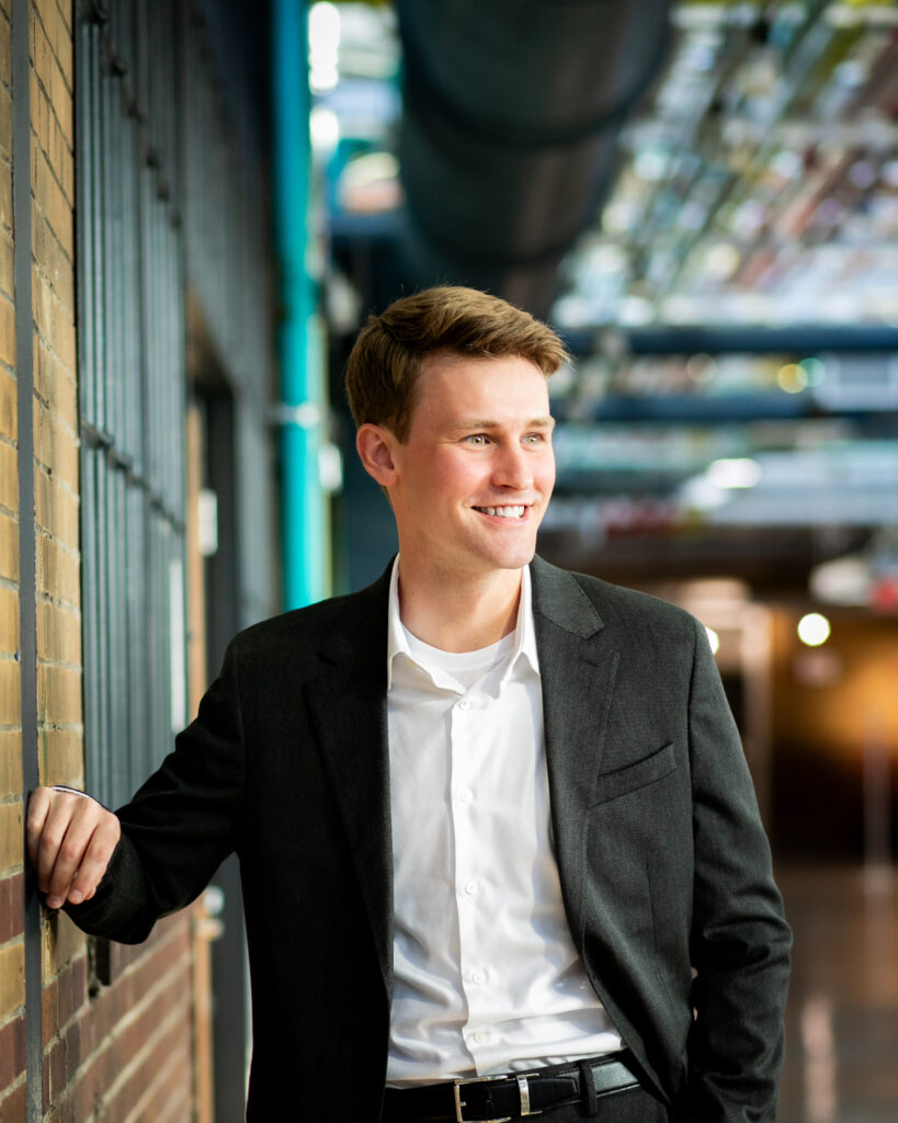 A man stands in a white button up shirt and a black suit jacket, leaning against a brick wall and looking off to the right.