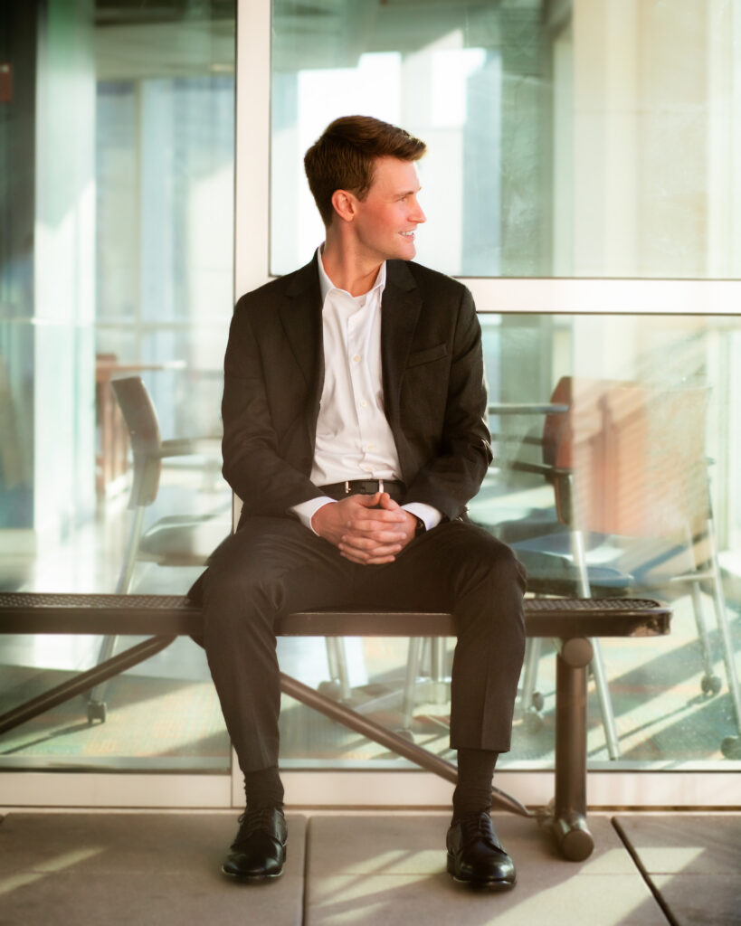 A man in a white shirt and suit jacket sits on a bench with his hands clasped in front of him and head looking off to the right.