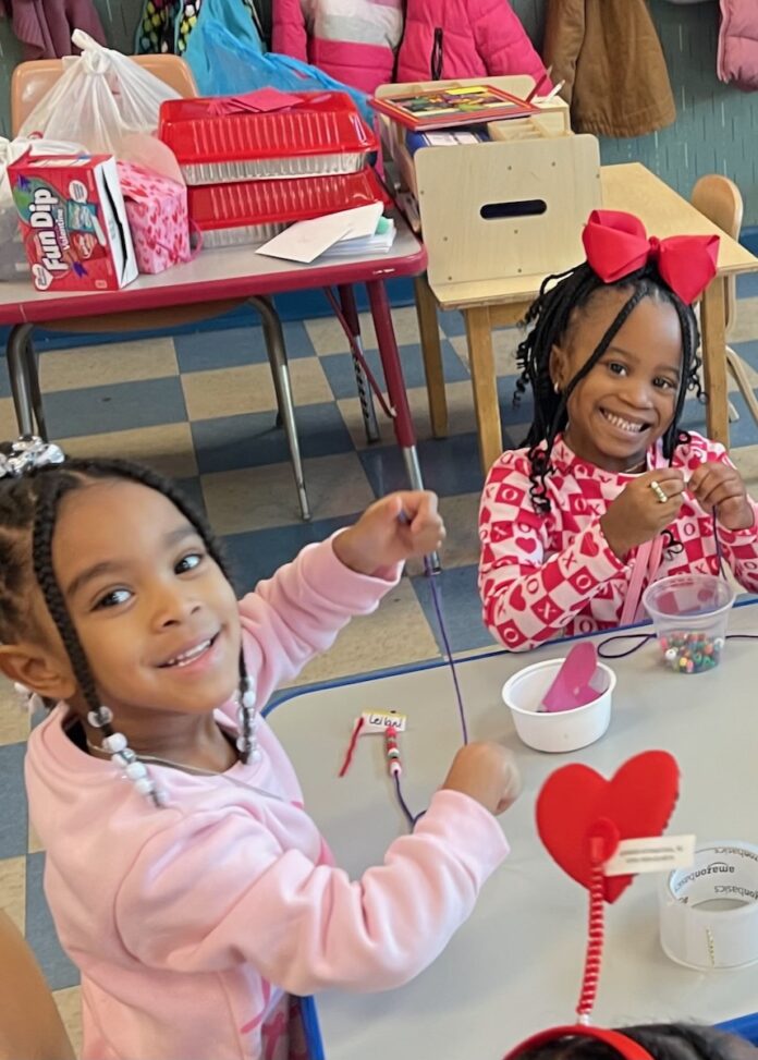 Two little girls in pink and red clothes make a Valentine's day craft at a table.
