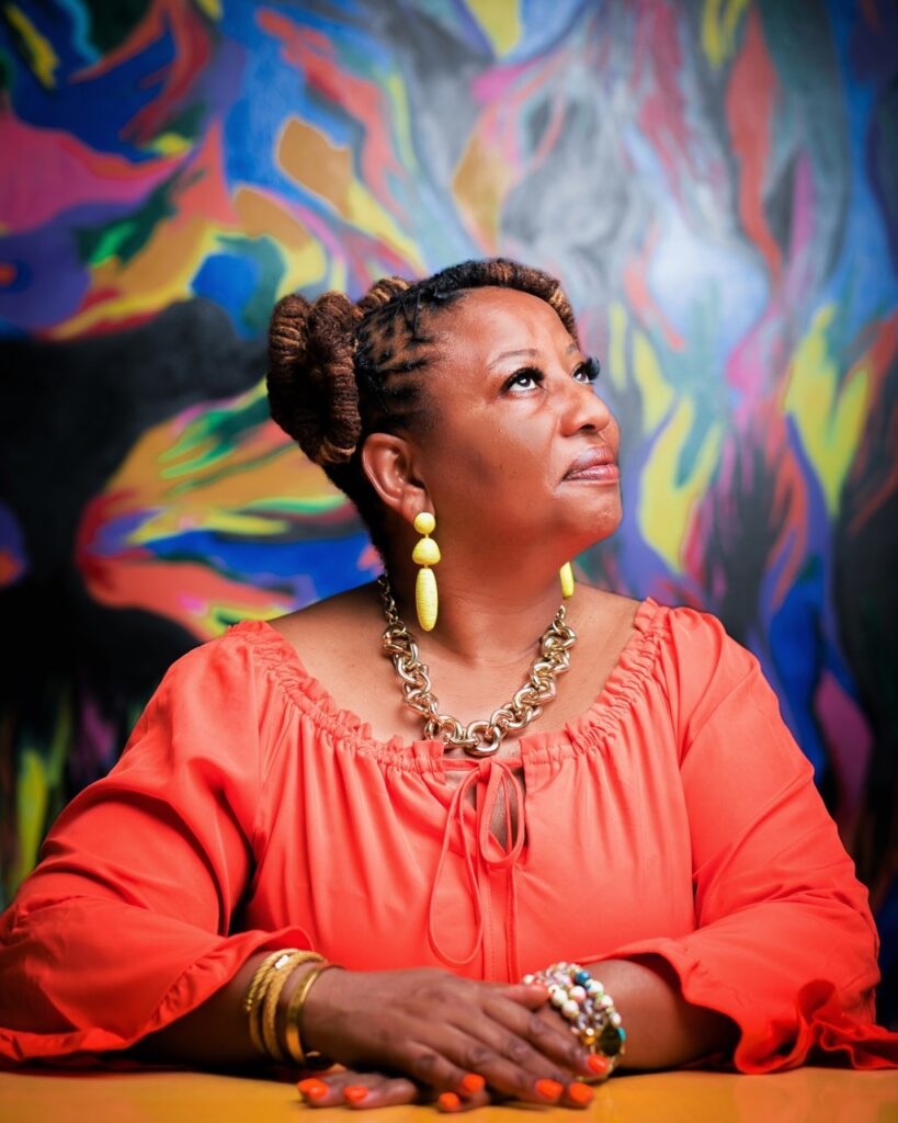 A woman in a coral long sleeve shirt from Rise Up 365 sits with her hands in front of her and a colorful wall behind her. 