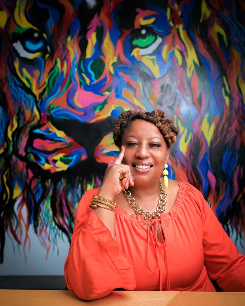 A woman sits at a wood table with her head resting on her hand and a rainbow lion painting behind her.