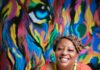 Rise Up 365 Works to Support the Youth of Pittsburgh A woman for Rise Up 365 sits in a coral long sleeve shirt in front of a rainbow lion painting.