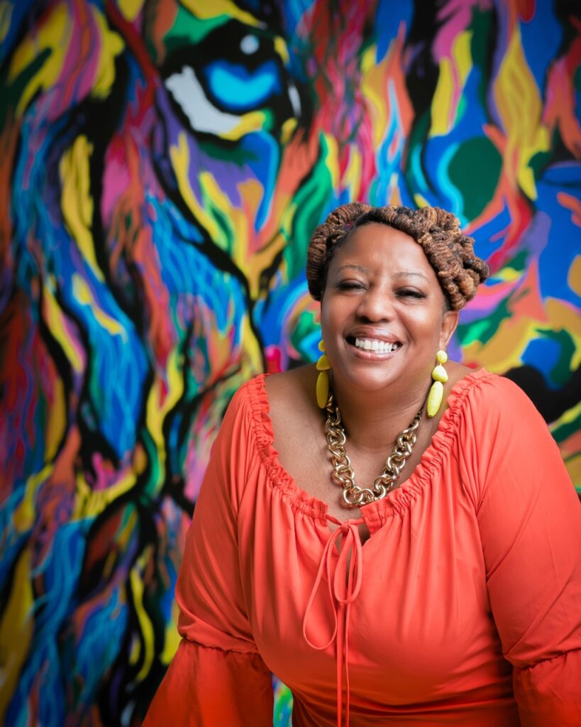 A woman for Rise Up 365 sits in a coral long sleeve shirt in front of a rainbow lion painting.