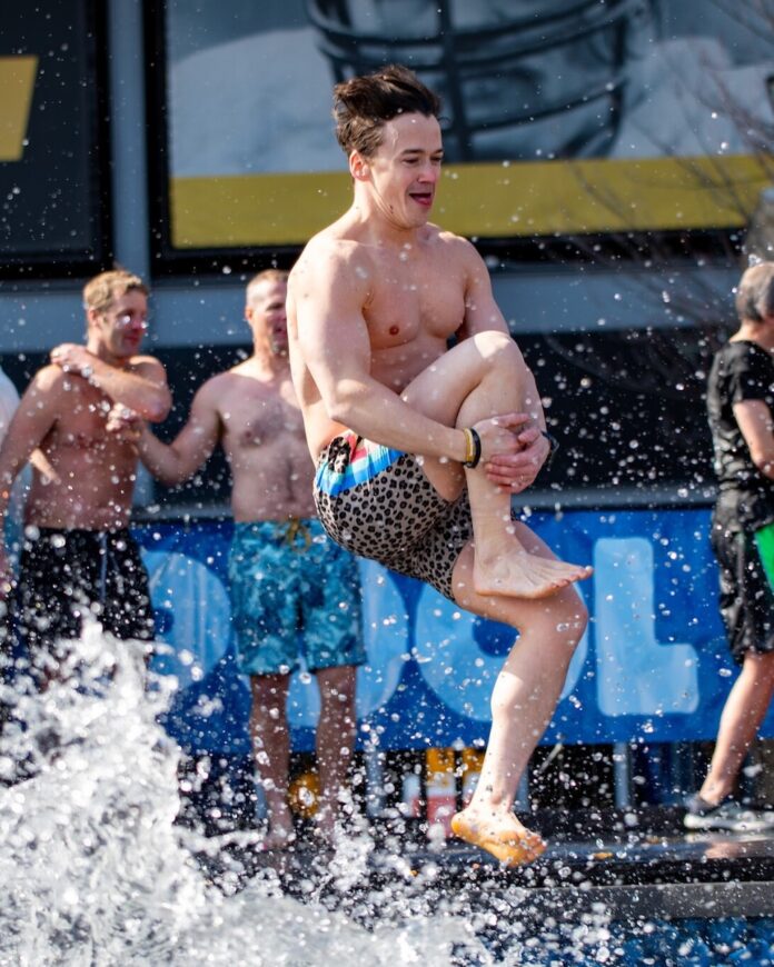 A man in blue swim shorts jumps into a pool while holding his knee.