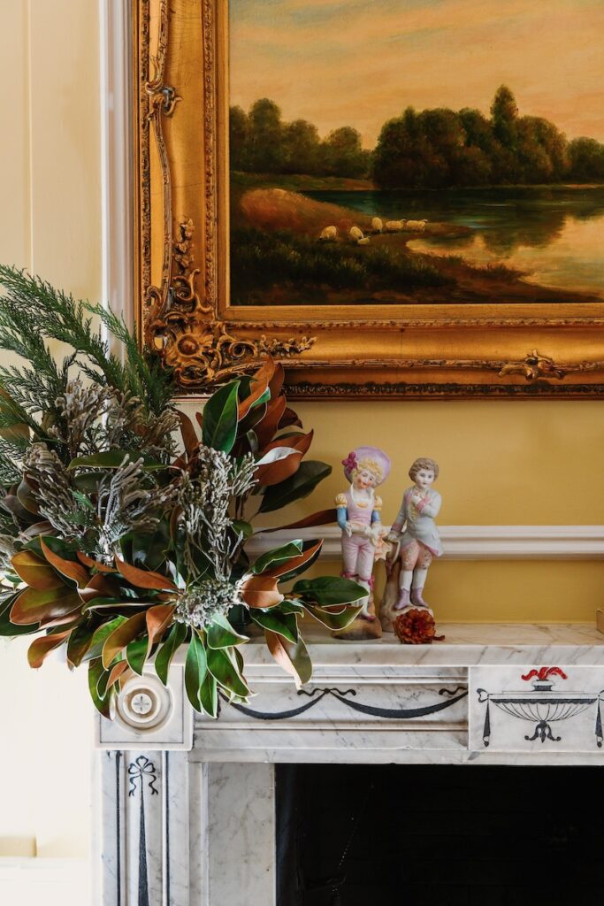 Two porcelain figures sit on a mantlepiece beside a landscape painting and a plant setting