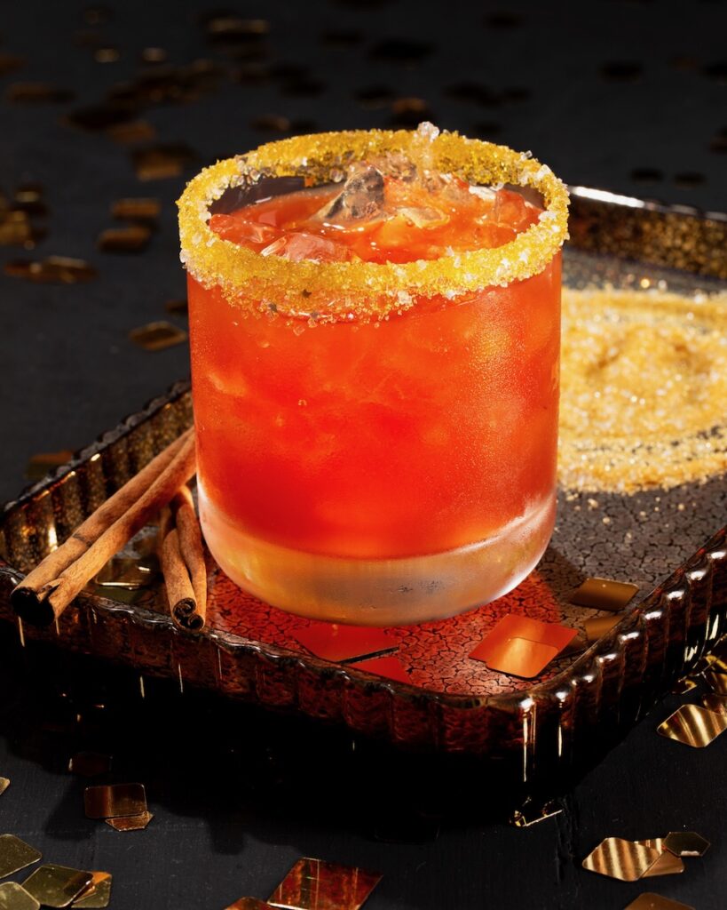 An orange cocktail with a sugar-dusted rim, surrounded by sticks of cinnamon 