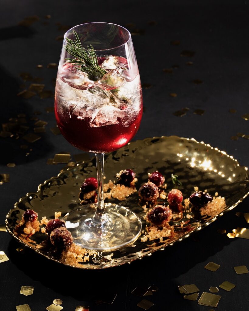 A cranberry rosemary spritz, a magenta red cocktail garnished with sprigs of rosemary, staged festively on a black background