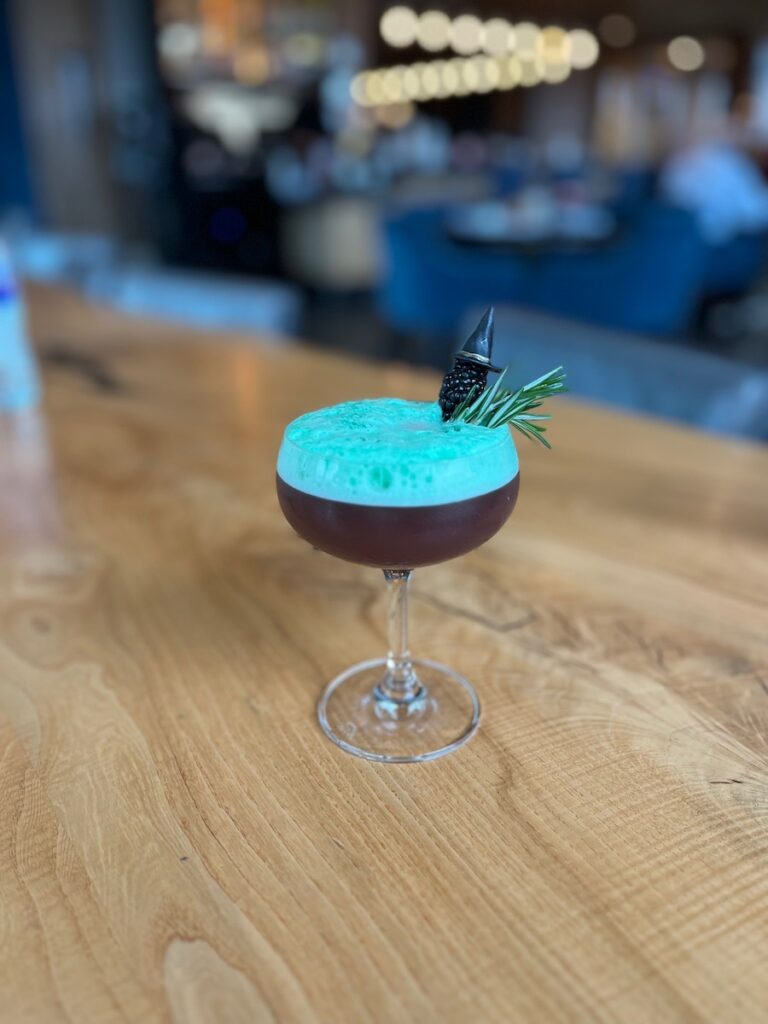 A turquoise and brown cocktail from Spirits & Tales with blackberry garnish with a small witch's hat