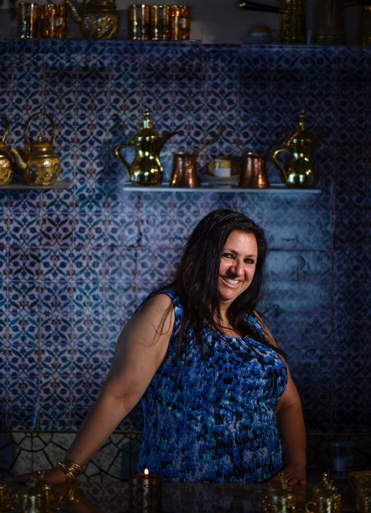 The owner of Khalil's, a smiling Middle Eastern woman in a blue shirt, poses for a camera