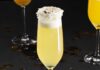 Look No Further Than Fine Wine & Good Spirits for New Year’s Cocktails A pale yellow drink in a champagne flute, garnished with coconut flakes on a black background