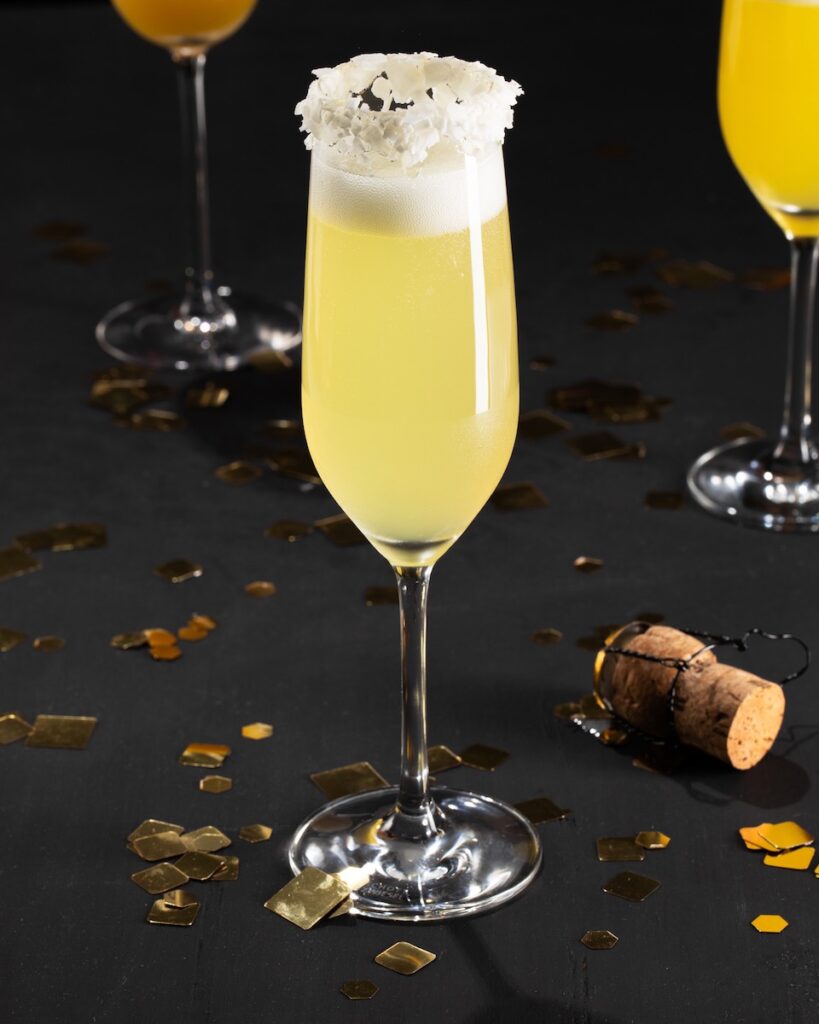 A pale yellow drink in a champagne flute, garnished with coconut flakes on a black background