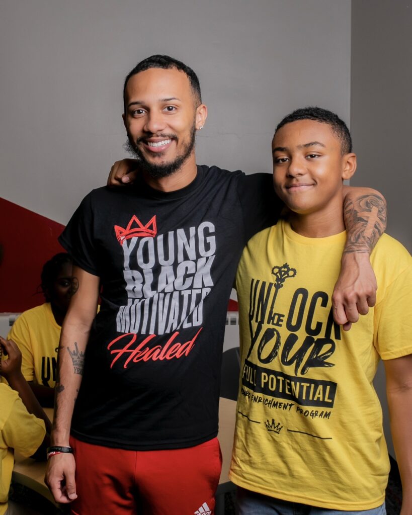 A man in a black Young Black Motivated Kings and Queens shirt stands with his arm around a kid in a yellow shirt. 