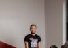 Guiding Pittsburgh’s Young Black Motivated Kings and Queens A man in a black Young Black Motivated Kings and Queens shirt stands at a wooden table that has four kids in yellow shirts seated at it, looking up at him.