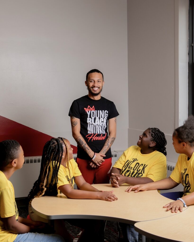 A man in a black Young Black Motivated Kings and Queens shirt stands at a wooden table that has four kids in yellow shirts seated at it, looking up at him.