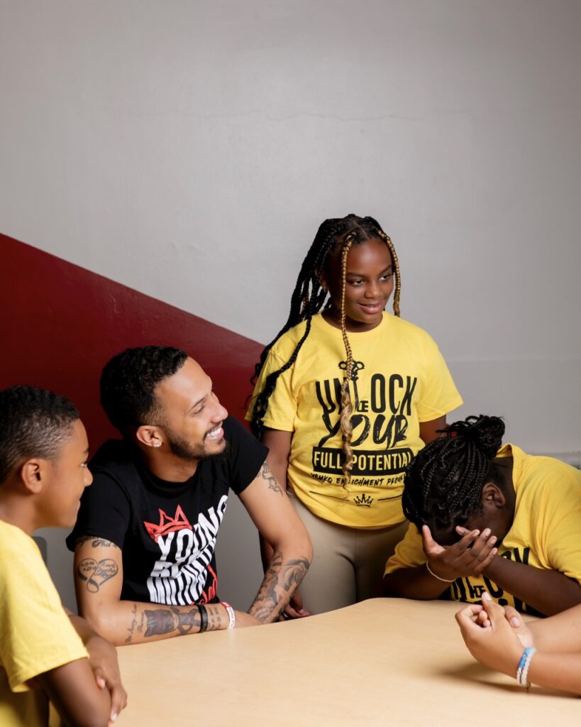 A man and three kids in yellow shirts laugh together at a wooden table.