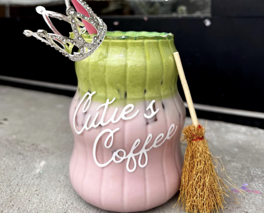 A pink and green strawberry matcha latte in a glass mug, staged with a broom beside it