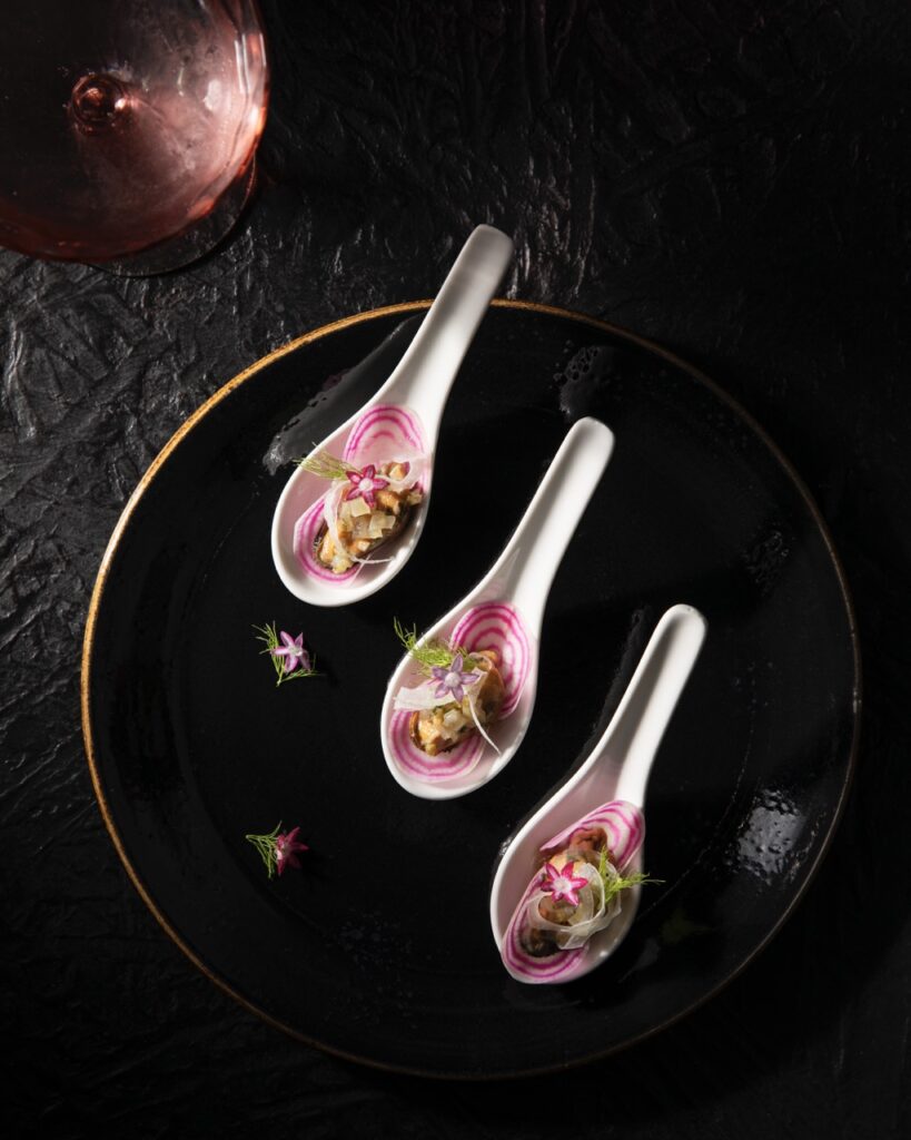 Three white spoons sit on a black plate holding slices of white and pink round vegetables with a pink edible flower on top.
