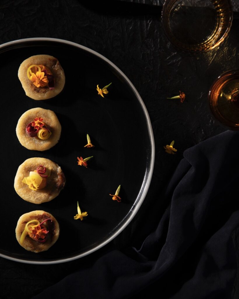 Four small round appetizer bites topped with yellow and red edible flowers sits on a black plate on a black background.