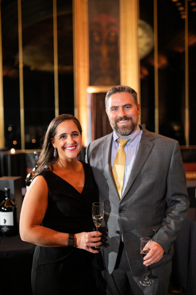 A man in a grey suit and yellow tie stands next to a woman in a black dress, both with wine glasses in hand.