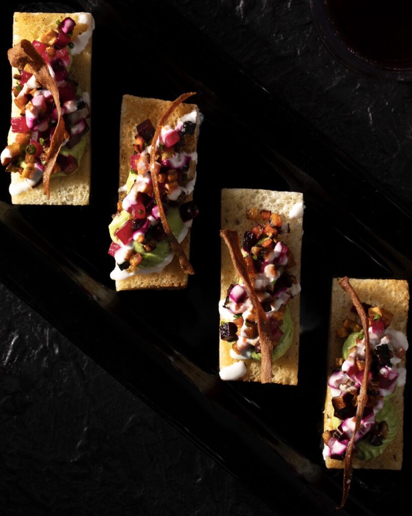 Four rectangular crackers sit on a black background with a salsa mix of vegetables topped on each.