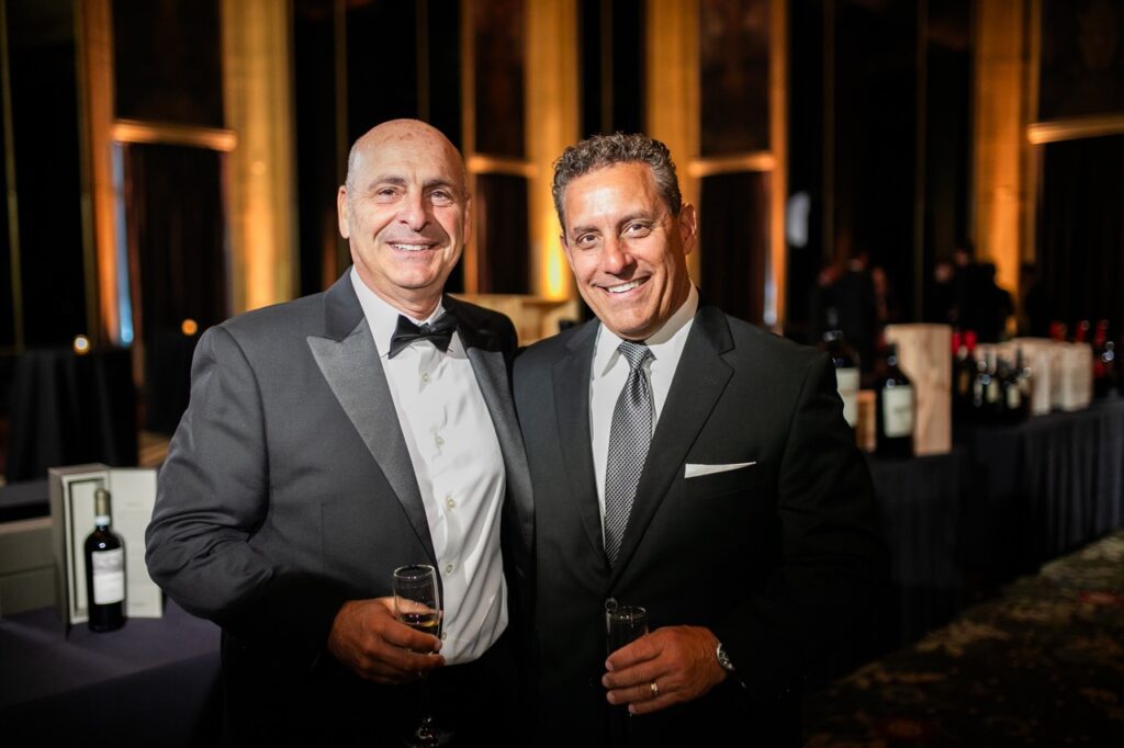 Two men in grey suits, one with a tie, one with a bowtie, stand with glasses of champagne in their hands.