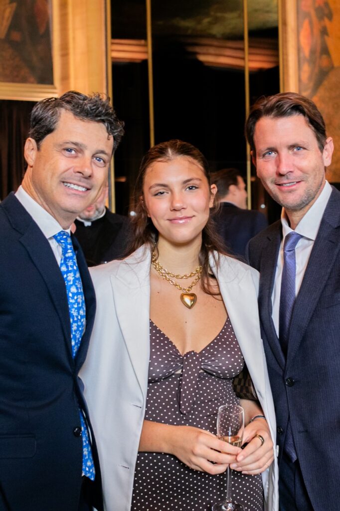 Two men in dark blue suits stand on both sides of a woman in a polka dot dress and a suit jacket over her shoulders.